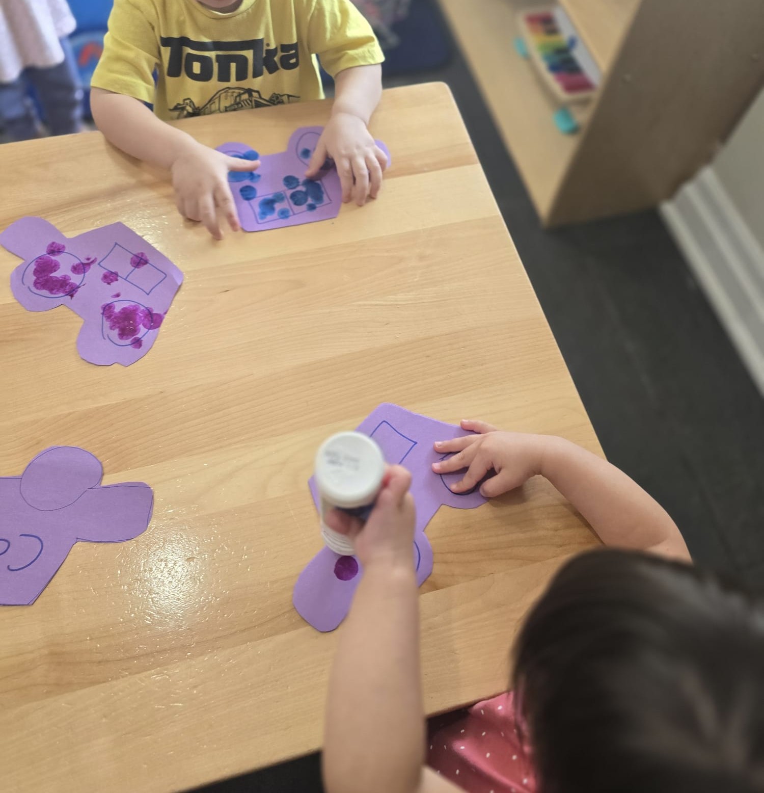 Toddlers stamping purple paint dots onto paper teddy bear cut-outs at a low wooden table