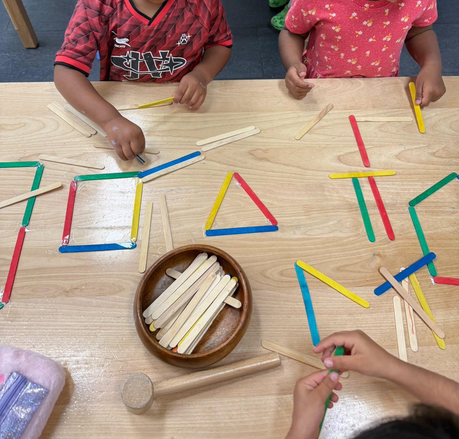 Children building squares and triangles from colourful craft sticks at a shared table