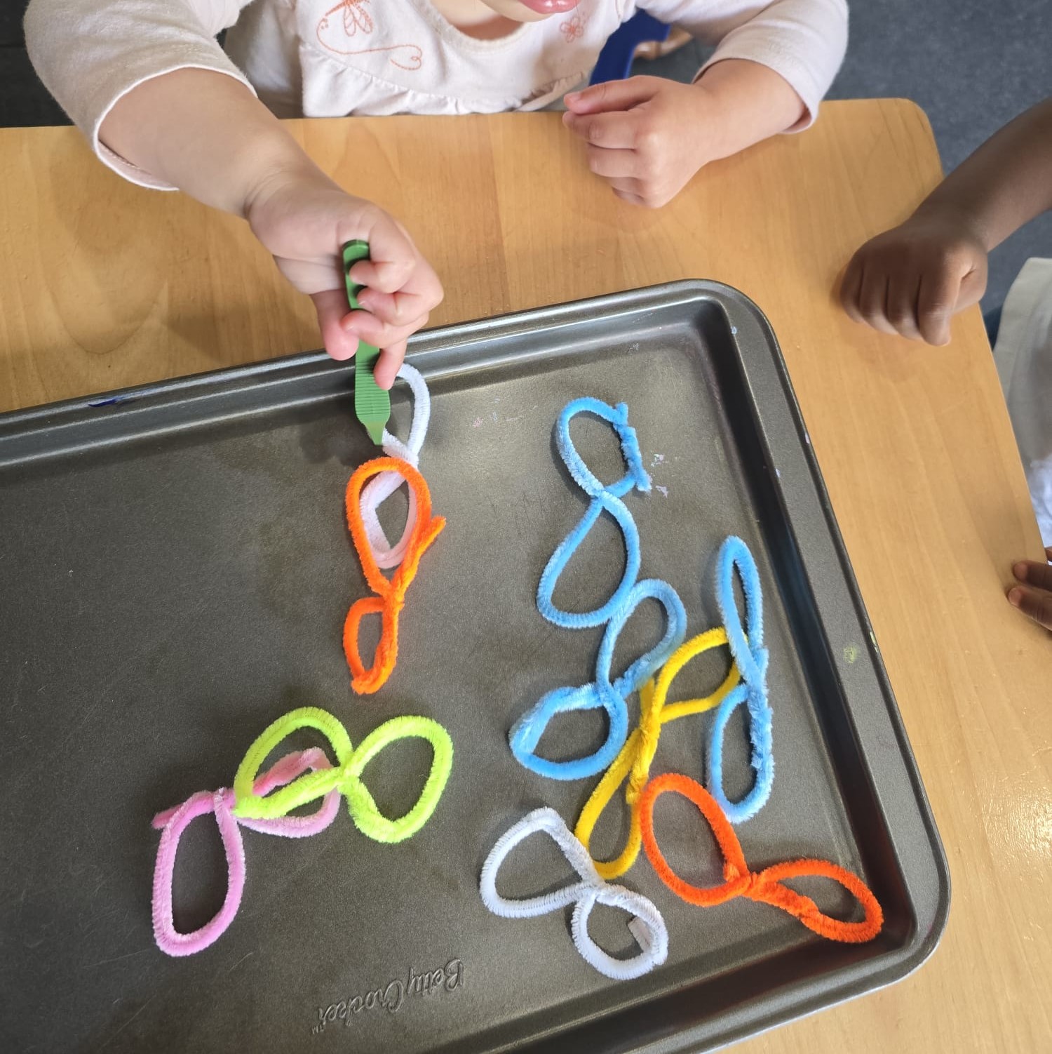 Child using tongs to pick up colourful pipe cleaner loops from a tray in a fine-motor activity