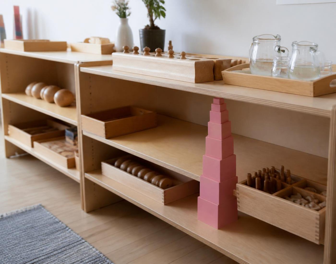 Low wooden Montessori shelves holding the pink tower, cylinder blocks and glass pouring pitchers