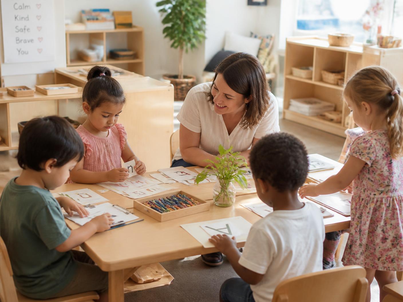 Educator sitting with a small group of nursery school children doing hands-on letter activities at a low wooden table
